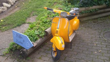 Gelbe Vespa-Roller steht auf einem Pflasterweg, umgeben von Grünpflanzen.